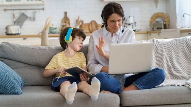 Hassled woman working on laptop on couch while child distracts her